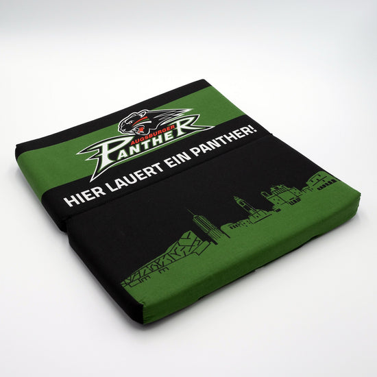 Faltbares schwarzes Sitzkissen mit grüner Augsburg-Skyline auf dem unteren Drittel "Hier lauert ein Panther!"-Schriftzug in der Mitte und Pantherlogo auf grünem Streifen im oberen Bereich.